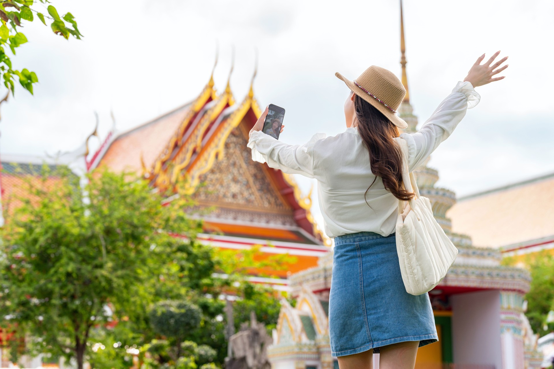 Rear View of Asian female tourist taking a selfie and video calling in front of Wat Phra Kaew and the Grand Palace, Bangkok, Thailand